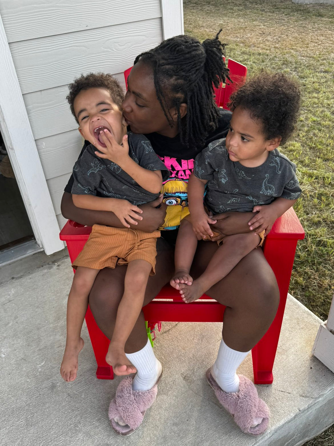 Woman sitting on a red chair with two children, one of whom is being kissed on the cheek.