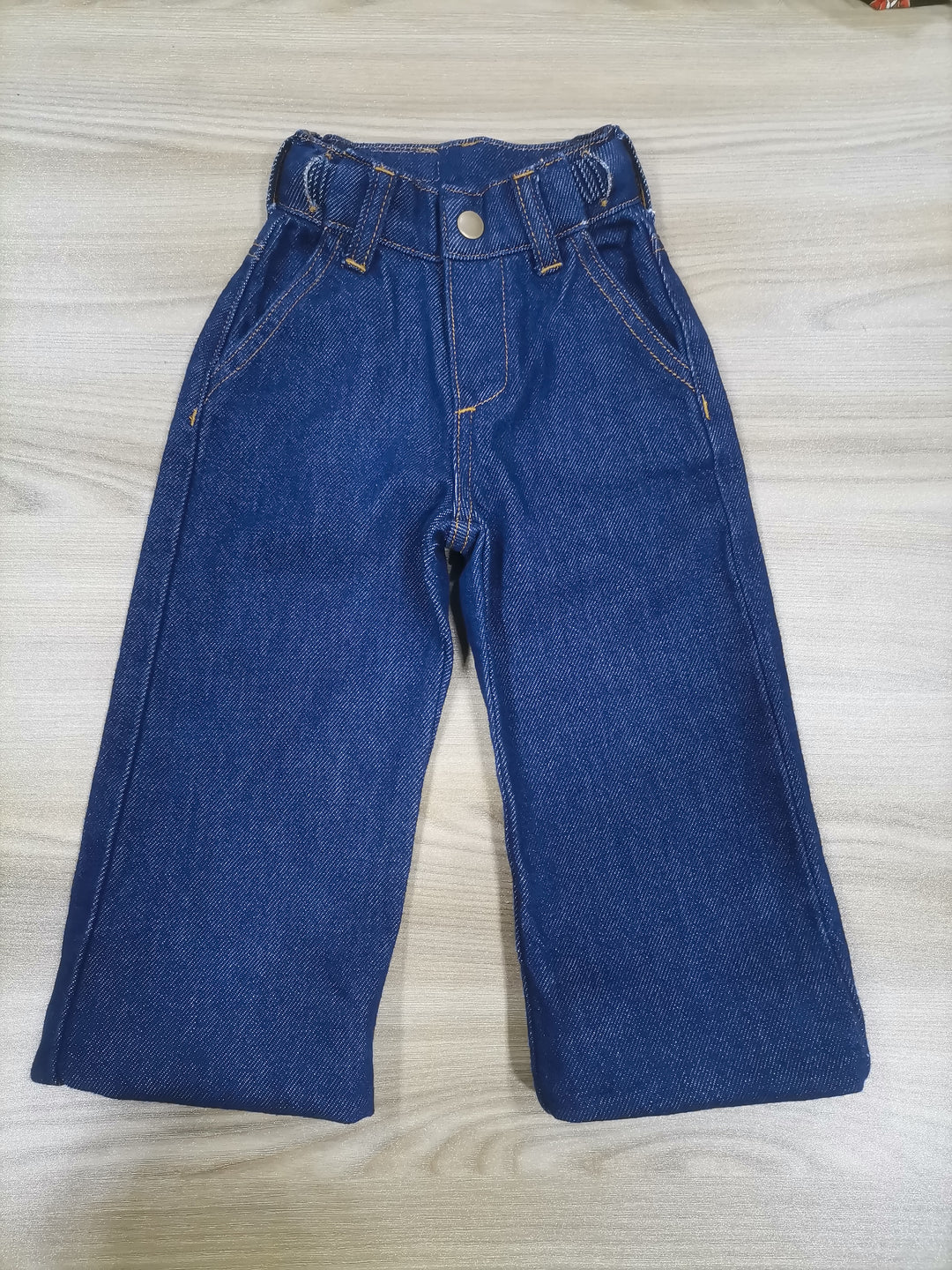 Front lay of a pair of blue denim jeans for kids laid flat on a wooden surface. Shortest length of extend hem expandable jeans